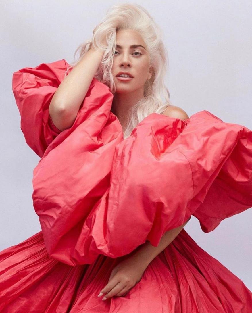Lady Gaga with voluminous platinum blonde hair wearing a bright red puffy dress against a plain light background
