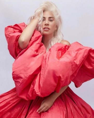 Lady Gaga with voluminous platinum blonde hair wearing a bright red puffy dress against a plain light background