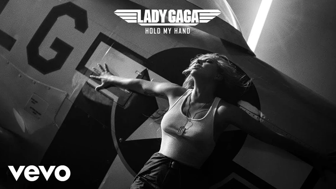 Black and white shot of Lady Gaga in a ribbed tank top next to a vintage fighter plane promoting her song, 'Hold My Hand.'