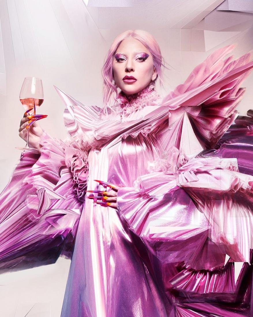 An adult female holding a glass of rose in a pink metallic dress with exaggerated shoulder ruffles, colorful manicure, and pink hair.