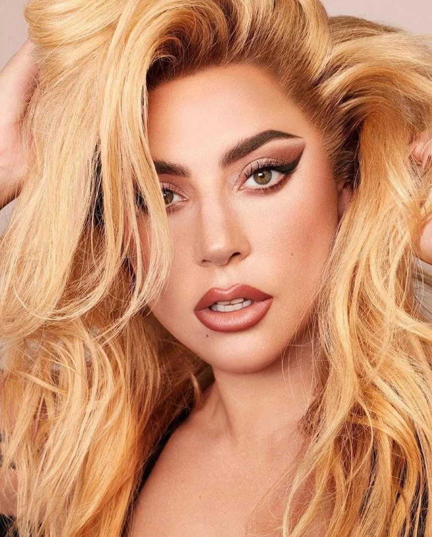 Close-up portrait of an adult female with blonde hair, dramatic makeup, and a neutral expression, posing with her hands in her hair.