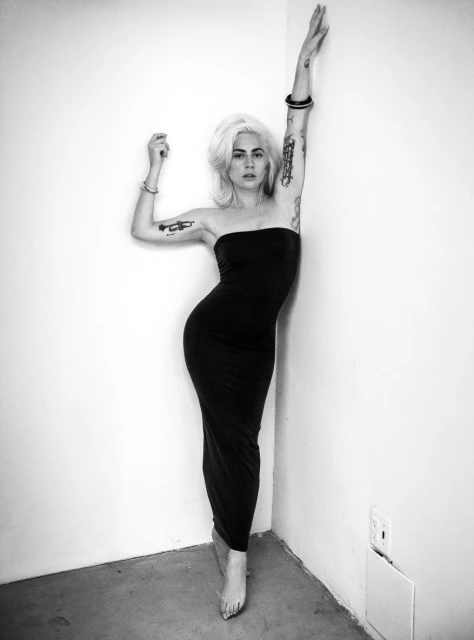 Black-and-white studio portrait of an adult woman with blonde hair, tattoos, and a black dress, posing near a corner wall.