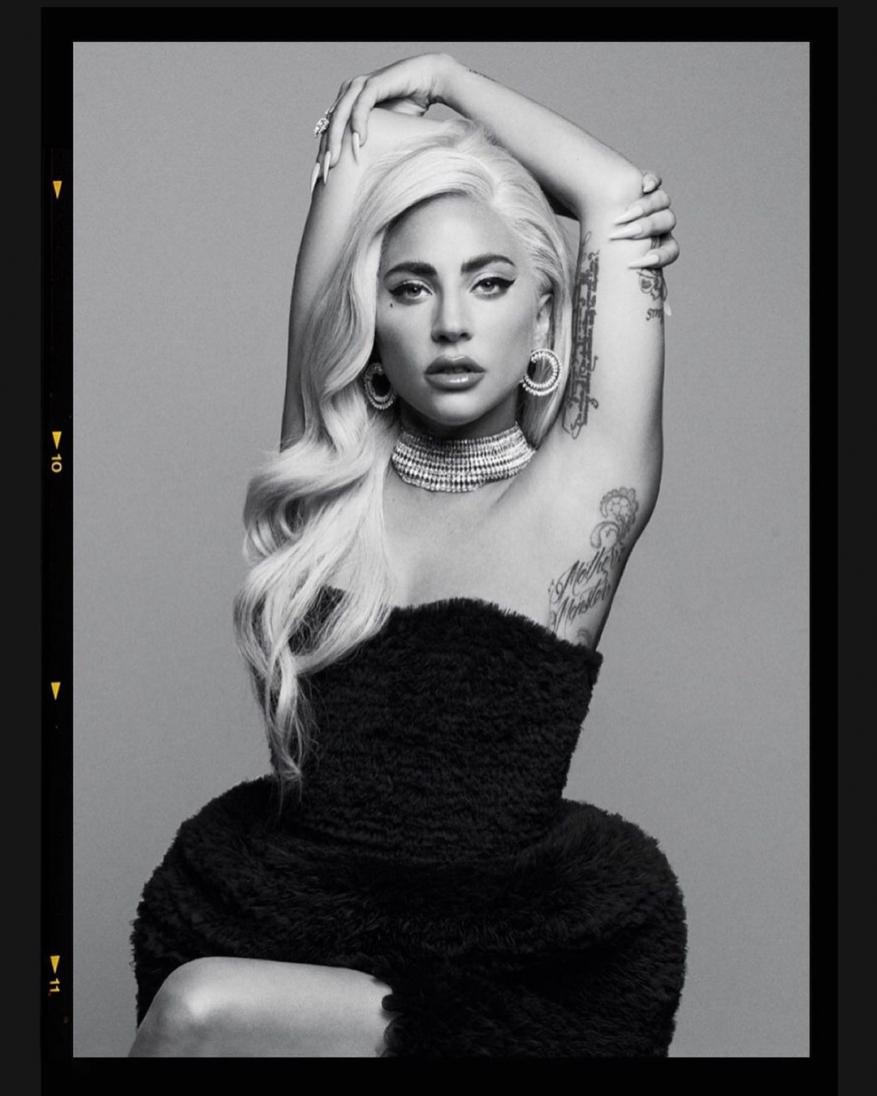 Monochrome portrait of a woman with blonde hair wearing a black strapless top and jewelry against a gray background.