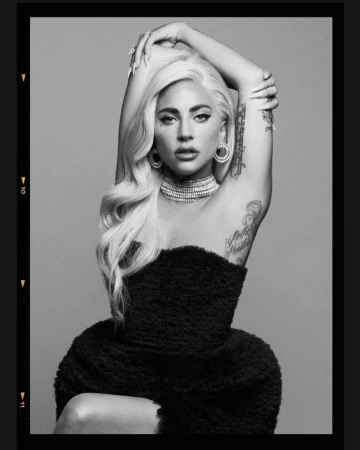Monochrome portrait of a woman with blonde hair wearing a black strapless top and jewelry against a gray background.