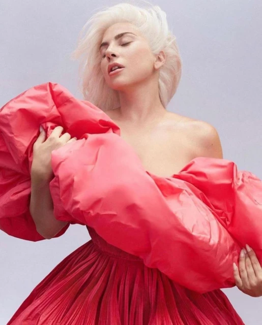 Woman with platinum blonde hair poses in a red, off-the-shoulder dress against a pale gray background in a medium portrait shot.