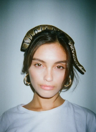 Portrait of a woman wearing a snake draped on her head as a crown with gold hoop earrings and a white t-shirt by Gabriel Perez Silva.