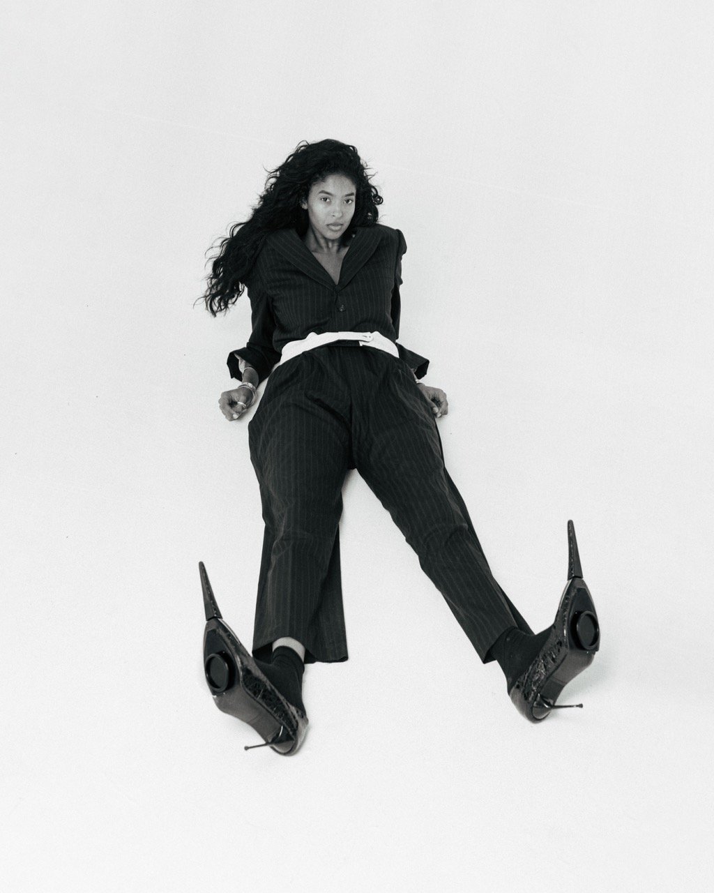 Elegant woman in black pinstripe suit and black heeled shoes sits on a white background. Fashion editorial shot.