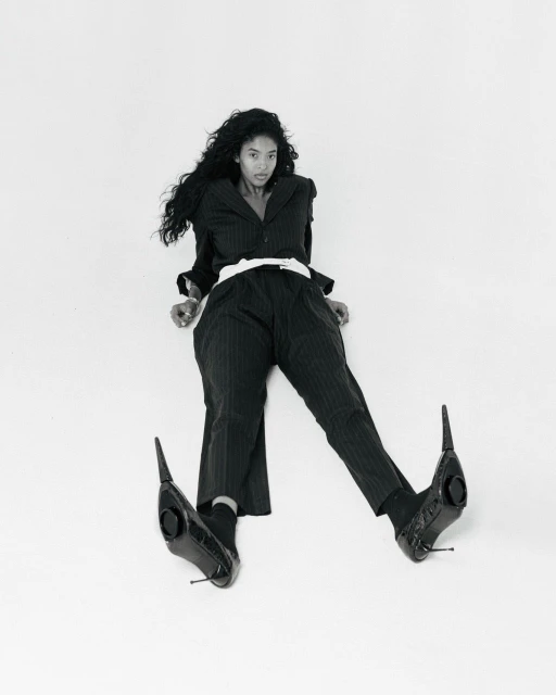 Elegant woman in black pinstripe suit and black heeled shoes sits on a white background. Fashion editorial shot.