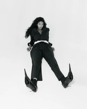 Elegant woman in black pinstripe suit and black heeled shoes sits on a white background. Fashion editorial shot.