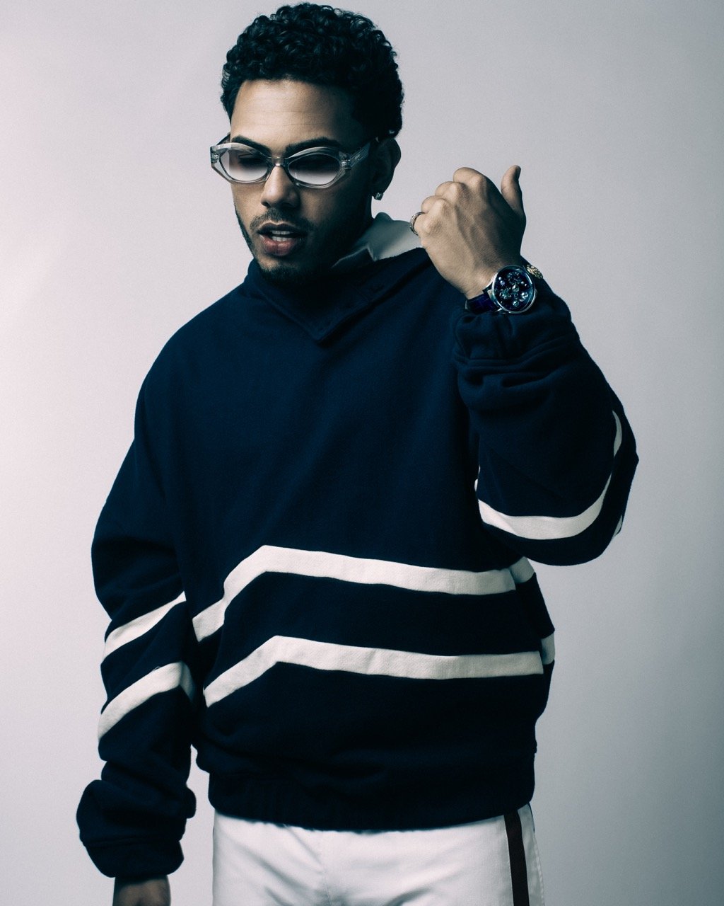 Studio shot of a young adult male wearing a navy and white sweater, glasses, and a watch, posing against a neutral background.