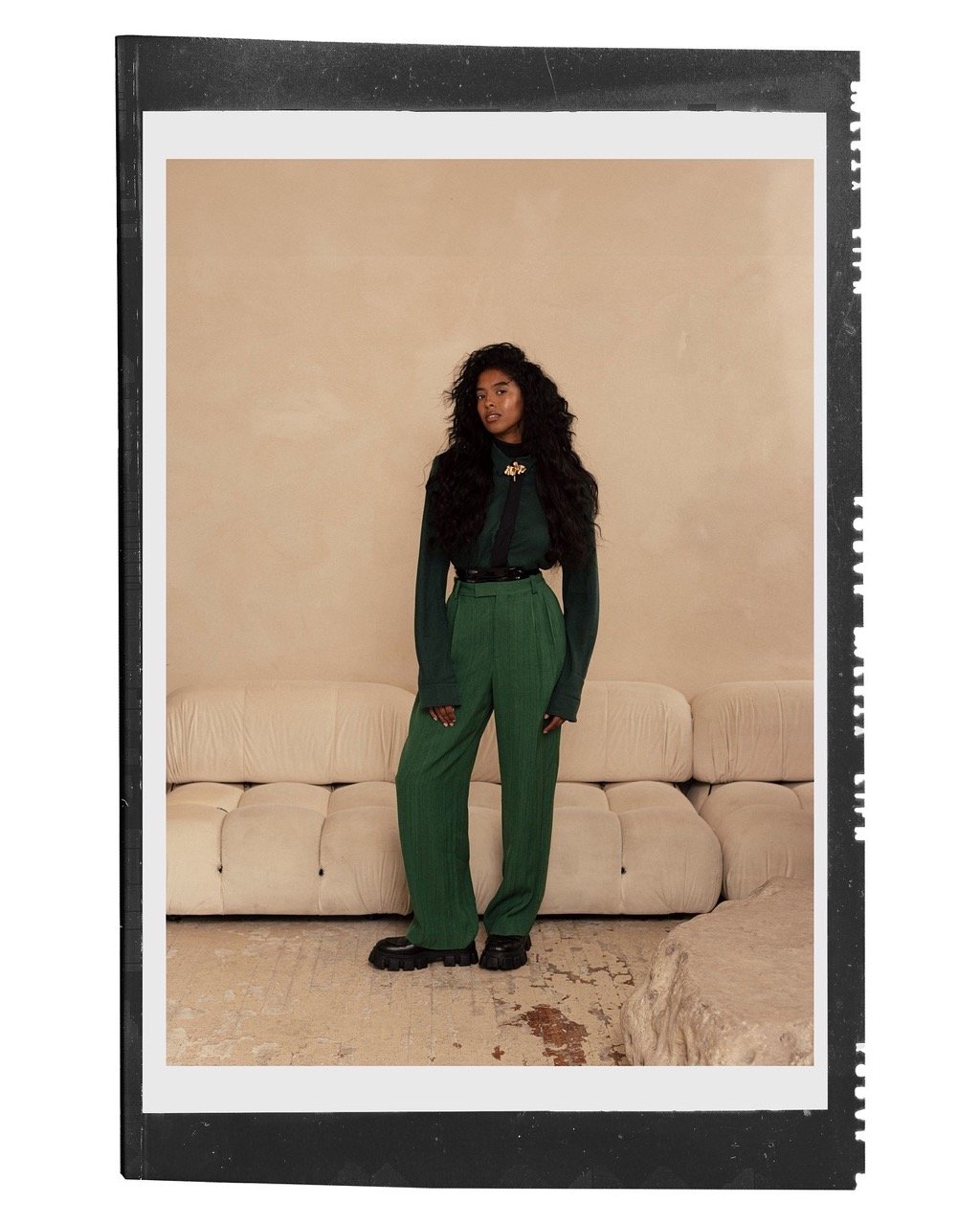 Full-body portrait of a woman with curly dark hair wearing a green sweater and pants in front of a light beige backdrop.