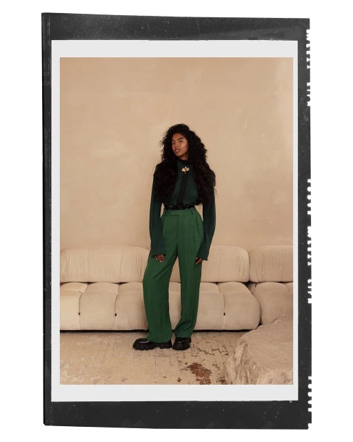 Full-body portrait of a woman with curly dark hair wearing a green sweater and pants in front of a light beige backdrop.