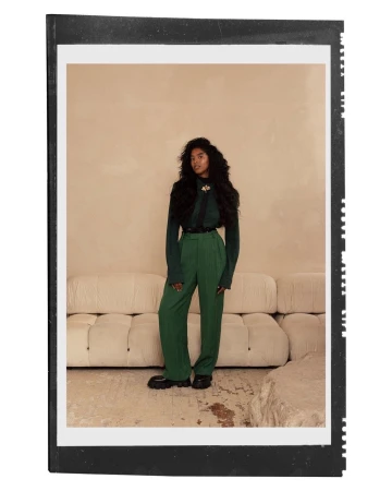 Full-body portrait of a woman with curly dark hair wearing a green sweater and pants in front of a light beige backdrop.