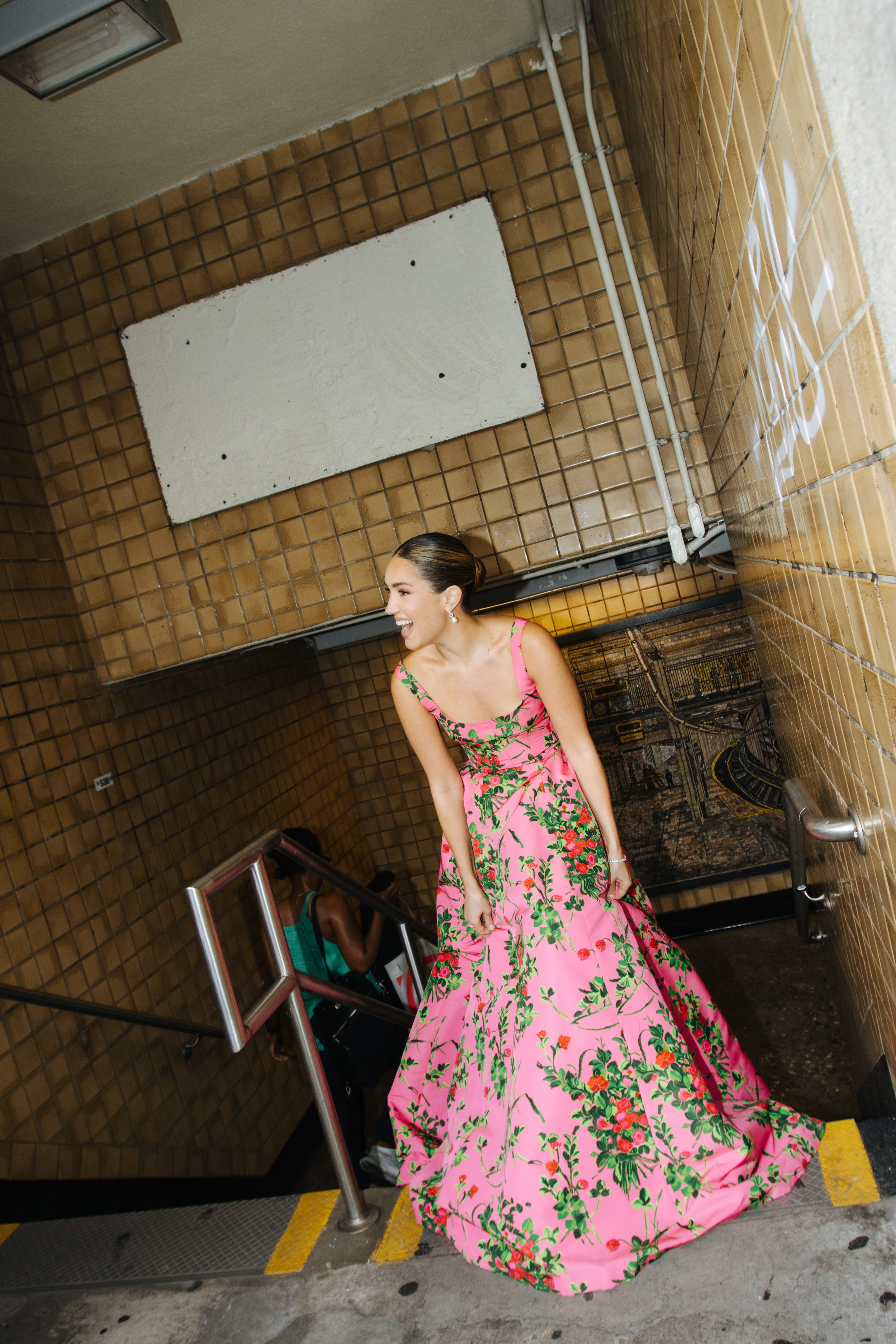 A woman in a pink floral gown descends stairs in a tiled, urban setting, captured with a tilted camera angle and a secondary female figure.