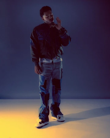 A man wearing a dark jacket and patchwork denim jeans poses in a studio with blue and yellow lighting.