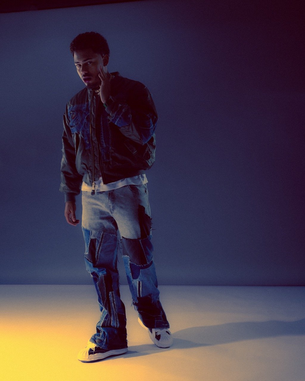 Young man in a bomber jacket and patchwork jeans poses in a photo studio with a grey background and bright yellow light.