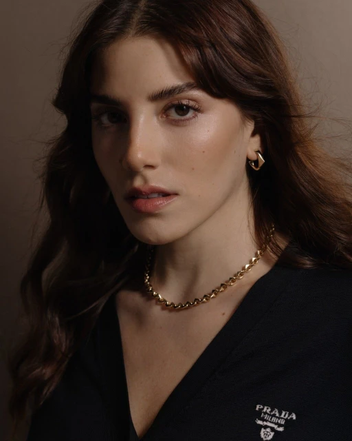 Close-up of a brunette woman with fair skin and neutral makeup, wearing a gold necklace and earrings in a neutral indoor setting.
