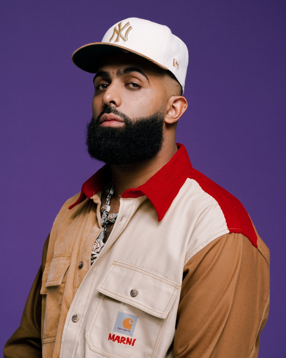 Portrait of an adult male with a beard wearing a baseball cap and a color-blocked jacket in front of a purple background.