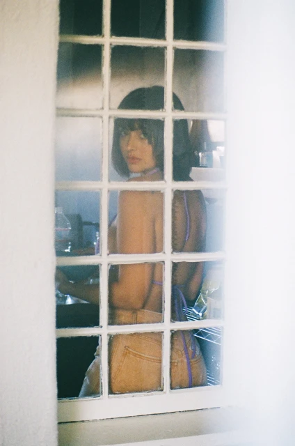 Indoor portrait of a young adult woman with dark hair posing in front of a window with white frames and a soft, hazy aesthetic.