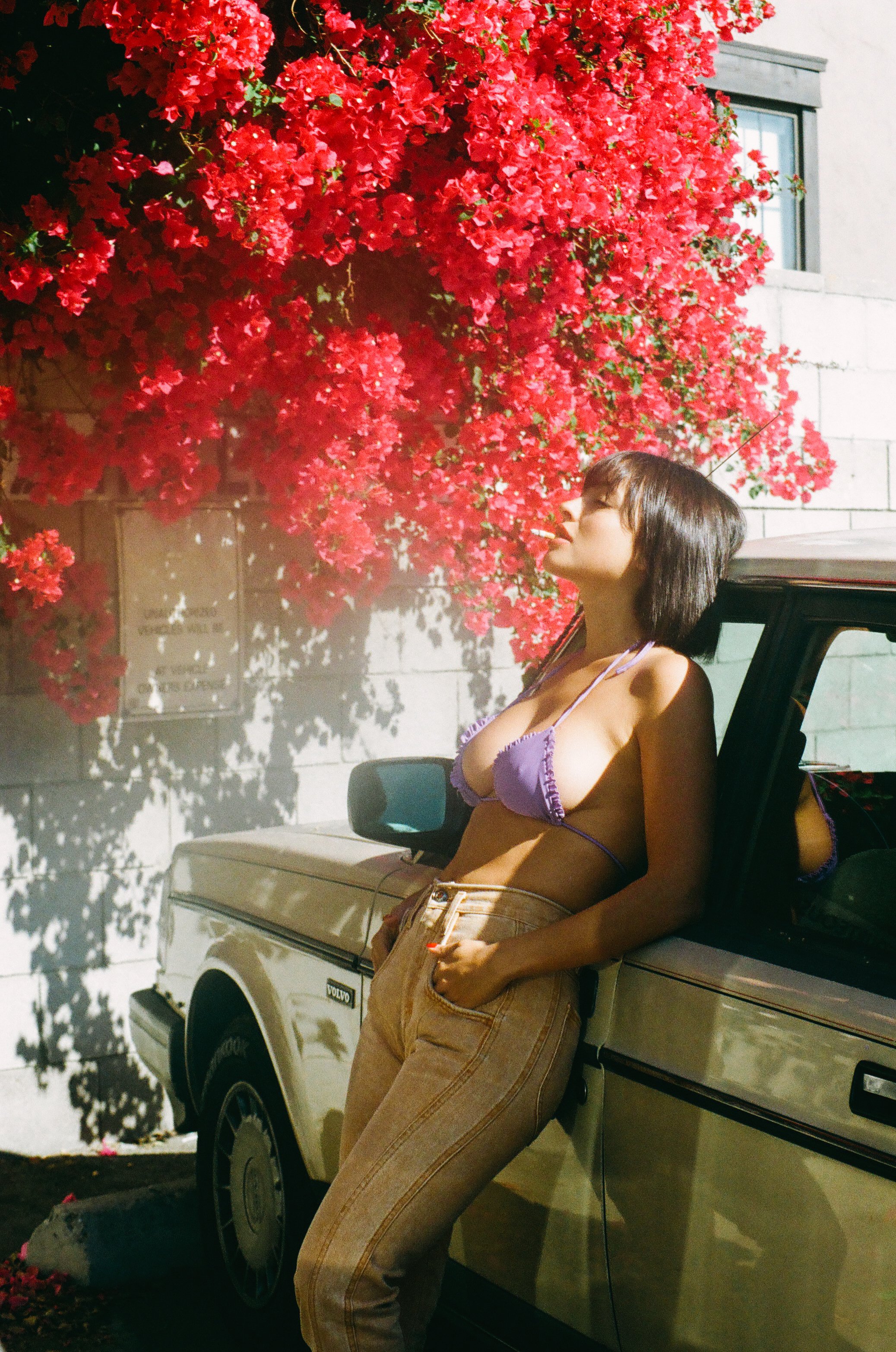Adult woman leans against car in a purple bikini top and tan pants, standing outdoors under a large bougainvillea bush on a sunny day.