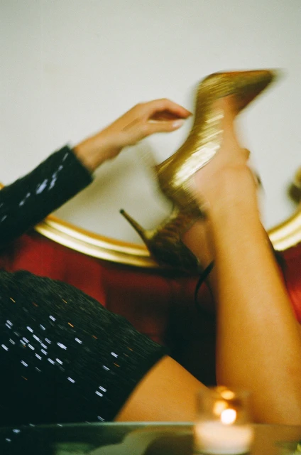 Woman wearing a sparkling black dress sitting on a red couch and holding up a gold, glittery stiletto heel in an indoor setting.
