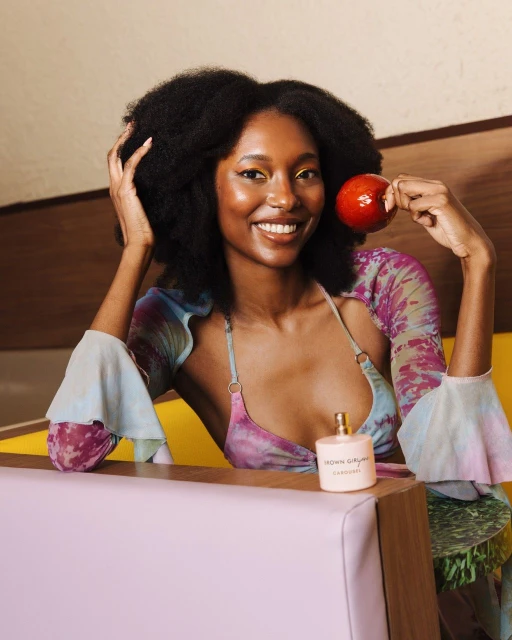 Smiling Black woman poses indoors, holding a red apple near a perfume bottle, wearing a tie-dye top with ruffled sleeves in soft, diffused light.