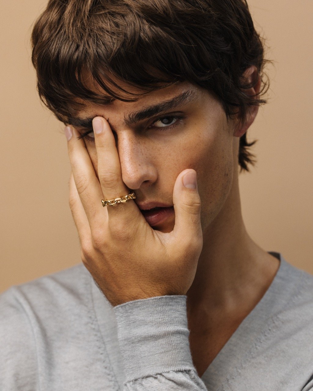 Close-up of a young adult male with brown hair wearing a gray sweater and a gold chain ring, posing indoors against a tan background.