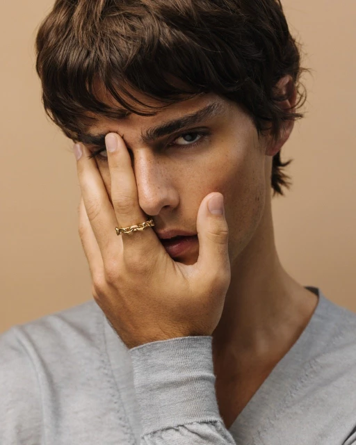 Close-up of a young adult male with brown hair wearing a gray sweater and a gold chain ring, posing indoors against a tan background.
