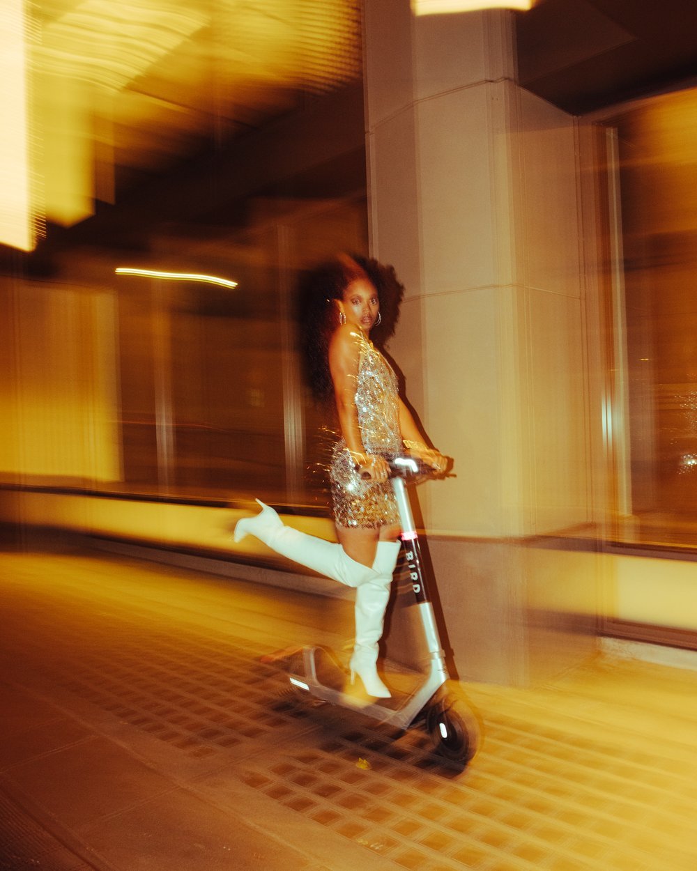 Woman in sparkling dress and white boots rides an electric scooter on a city sidewalk at night, illuminated by yellow lighting.