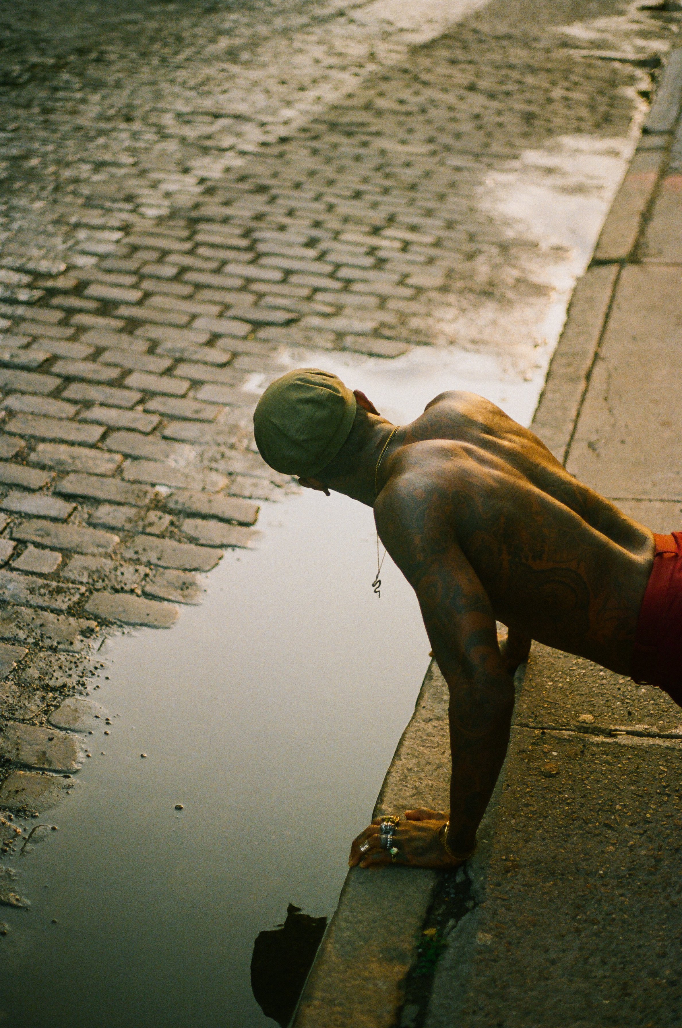 Tattooed man in a cap is doing push-ups near a puddle on a brick sidewalk, showcasing an urban fitness scene in diffused sunlight.