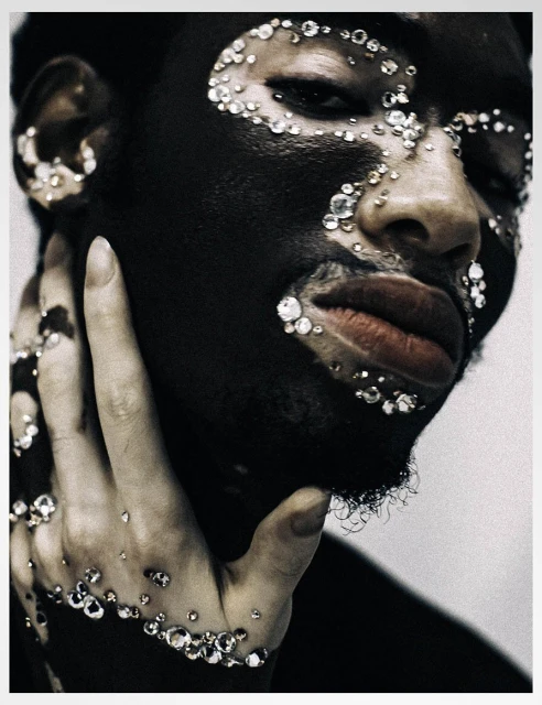 Close-up studio portrait of a Black adult with black body paint and sparkling jewels on face and hand against a white background.
