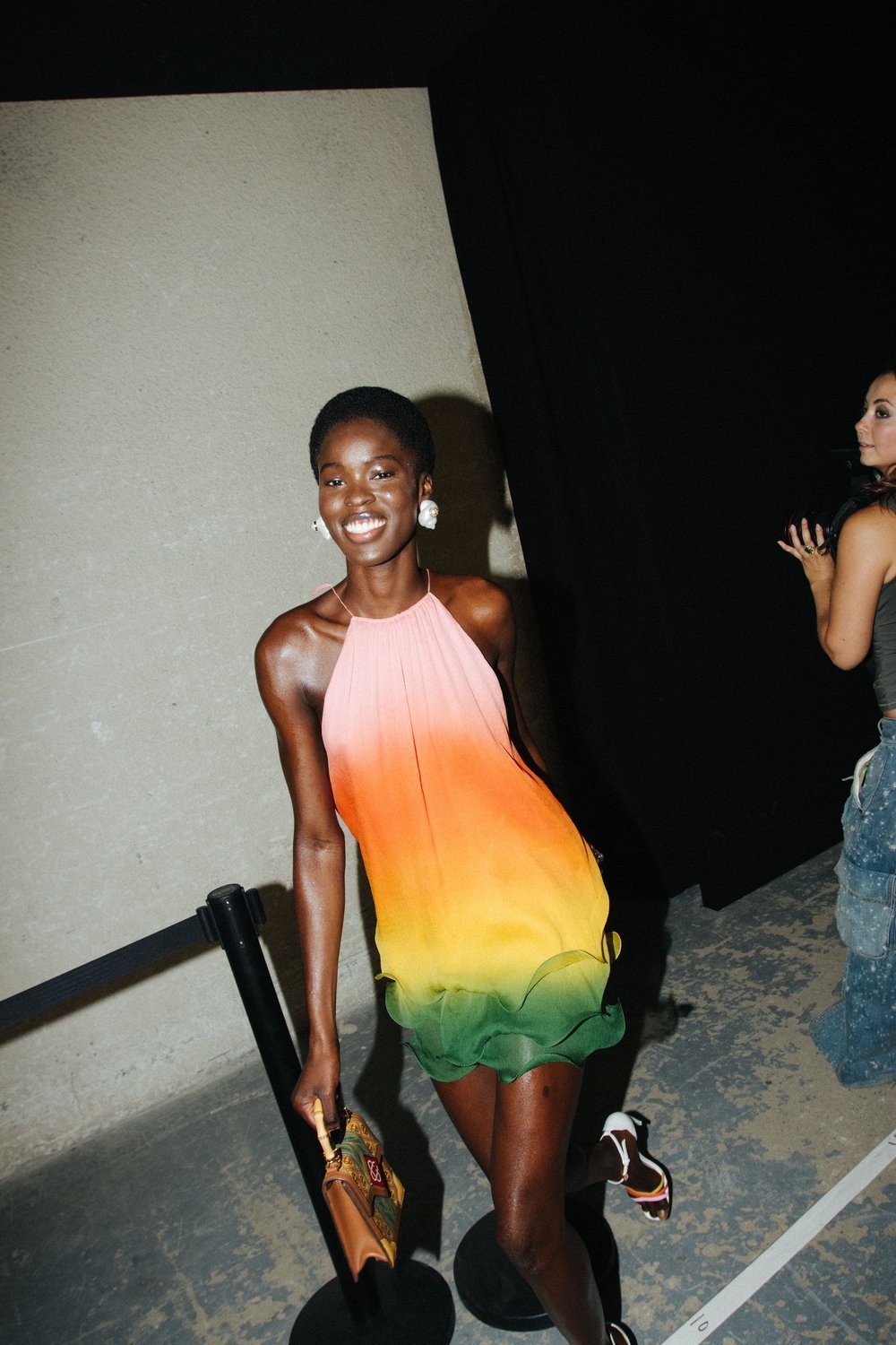 A smiling Black woman poses in an ombre minidress with a handbag, wearing white heeled sandals in an indoor environment.