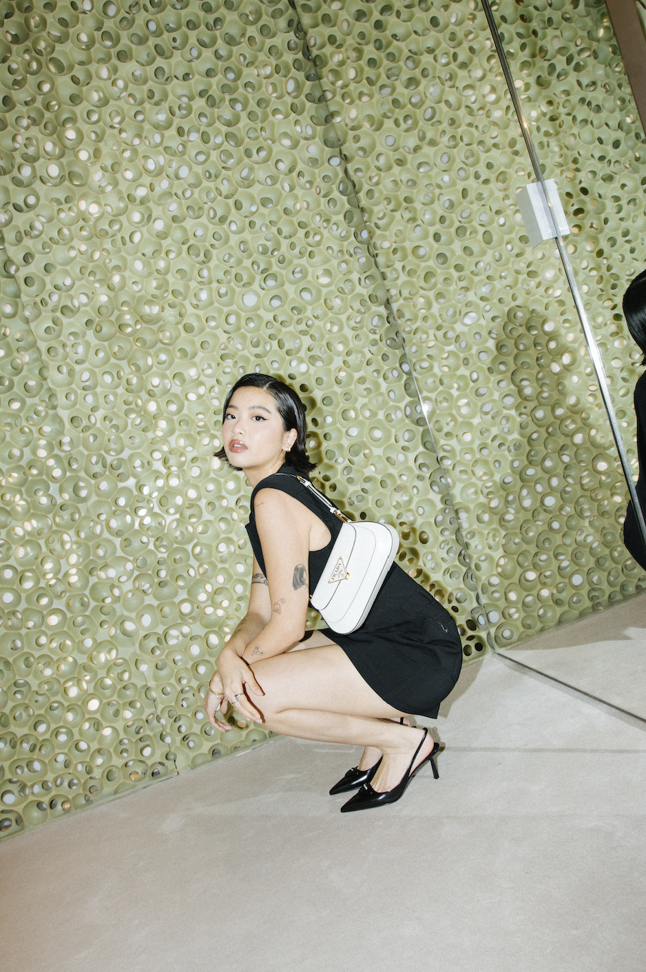 Woman in black dress poses in a crouched position with a white Prada bag against a decorative green wall, looking at the camera.