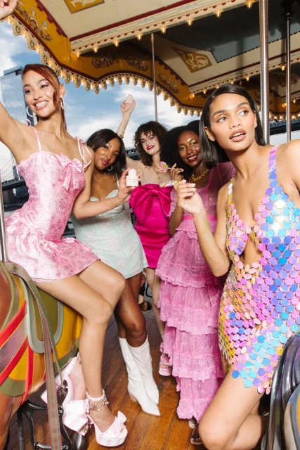 Five diverse women in pink dresses pose on a carousel in an outdoor city setting, showcasing fashion and beauty, styled for a special occasion.
