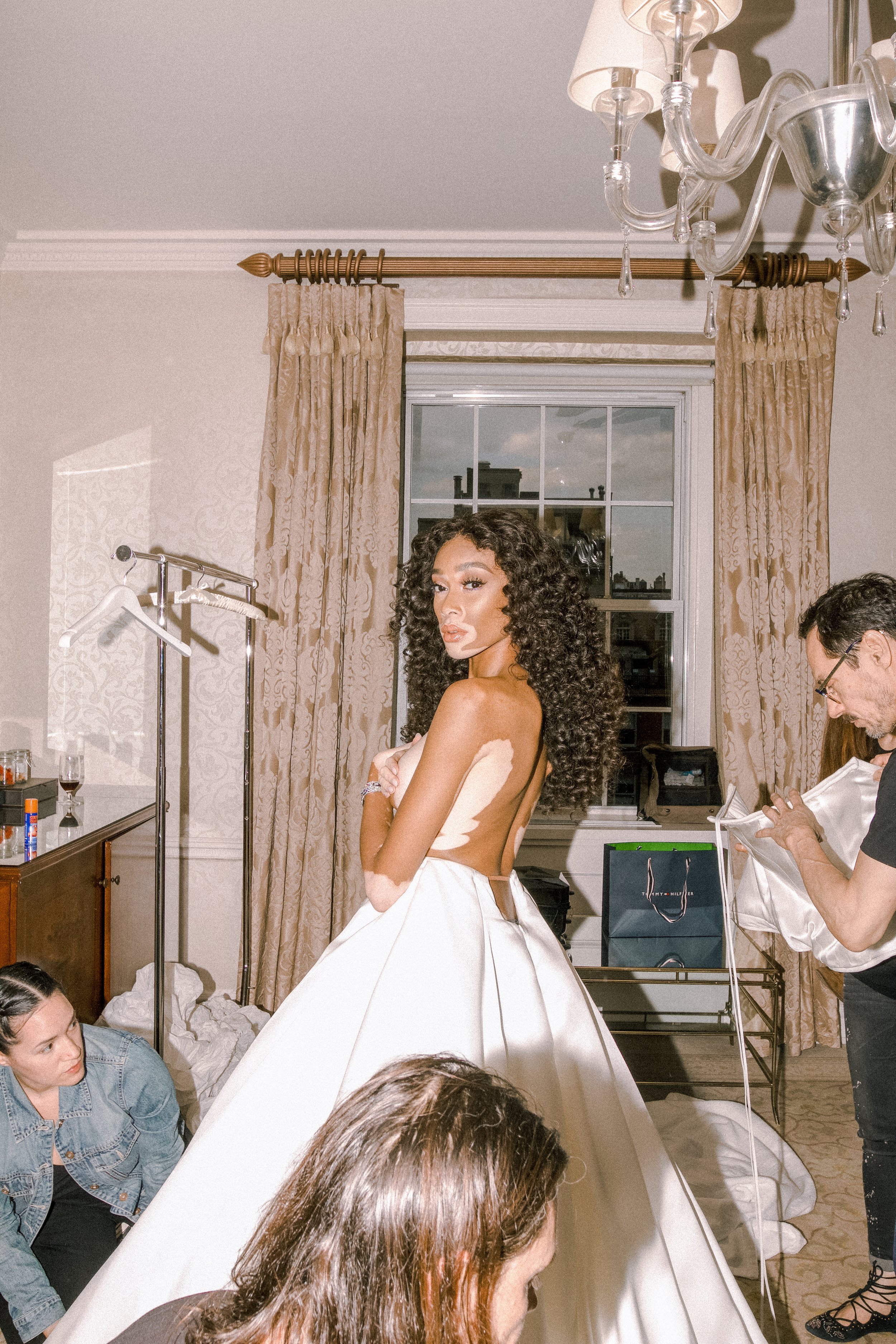 Winnie Harlow in a white wedding gown, posing in a room with stylists. Window, clothing rack, and formalwear bags are present.