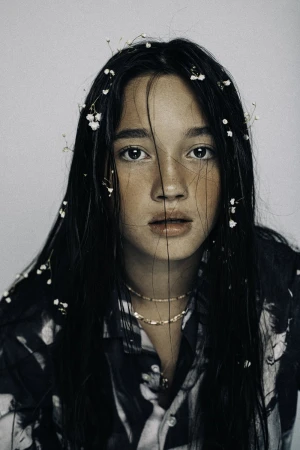 Close-up portrait of a young woman with dark wet hair adorned with tiny white flowers, wearing a patterned shirt and delicate necklaces.