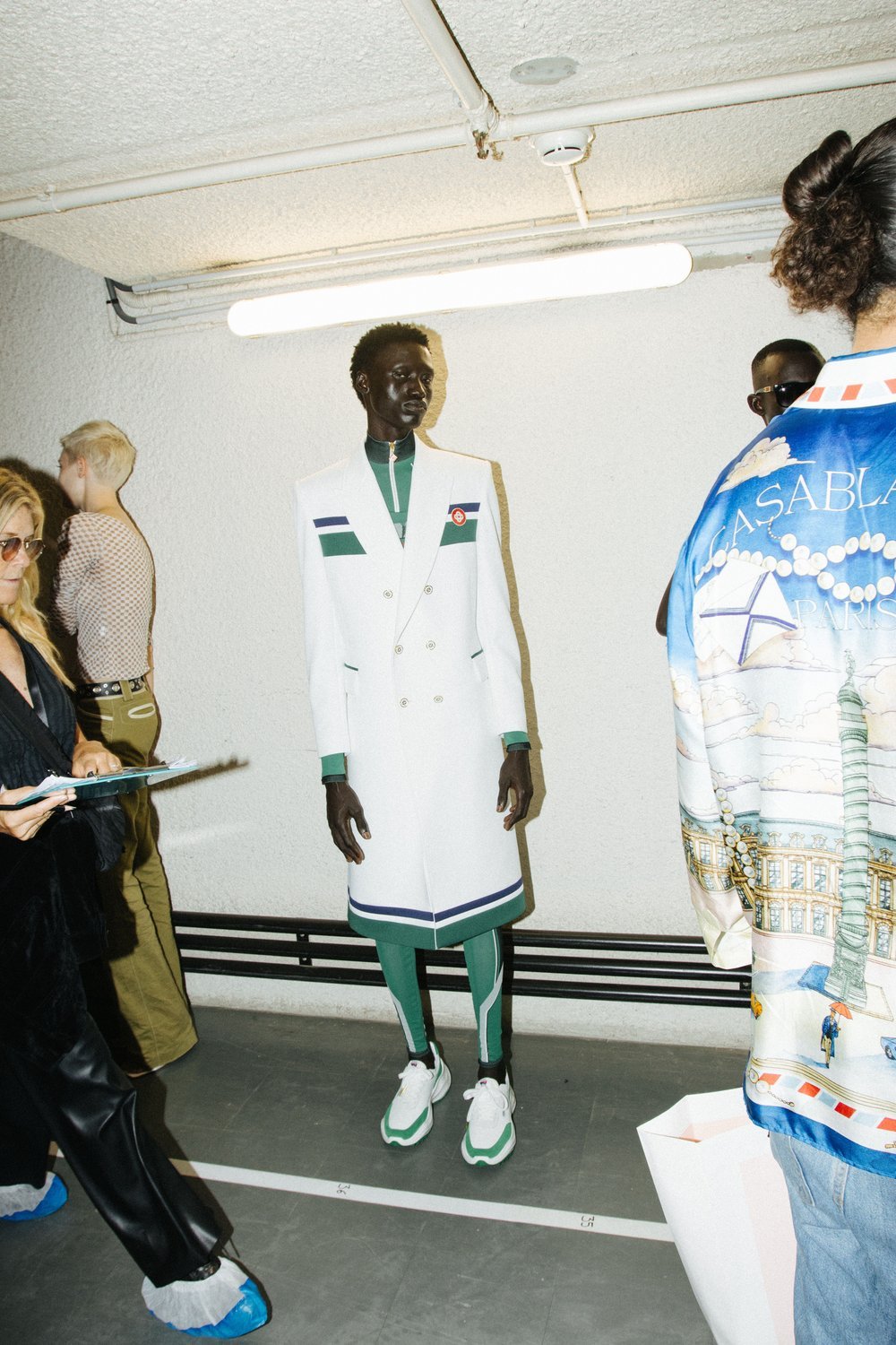 Full shot of a Black male model posing in a white coat with green trim, green leggings, and white sneakers in a backstage setting.