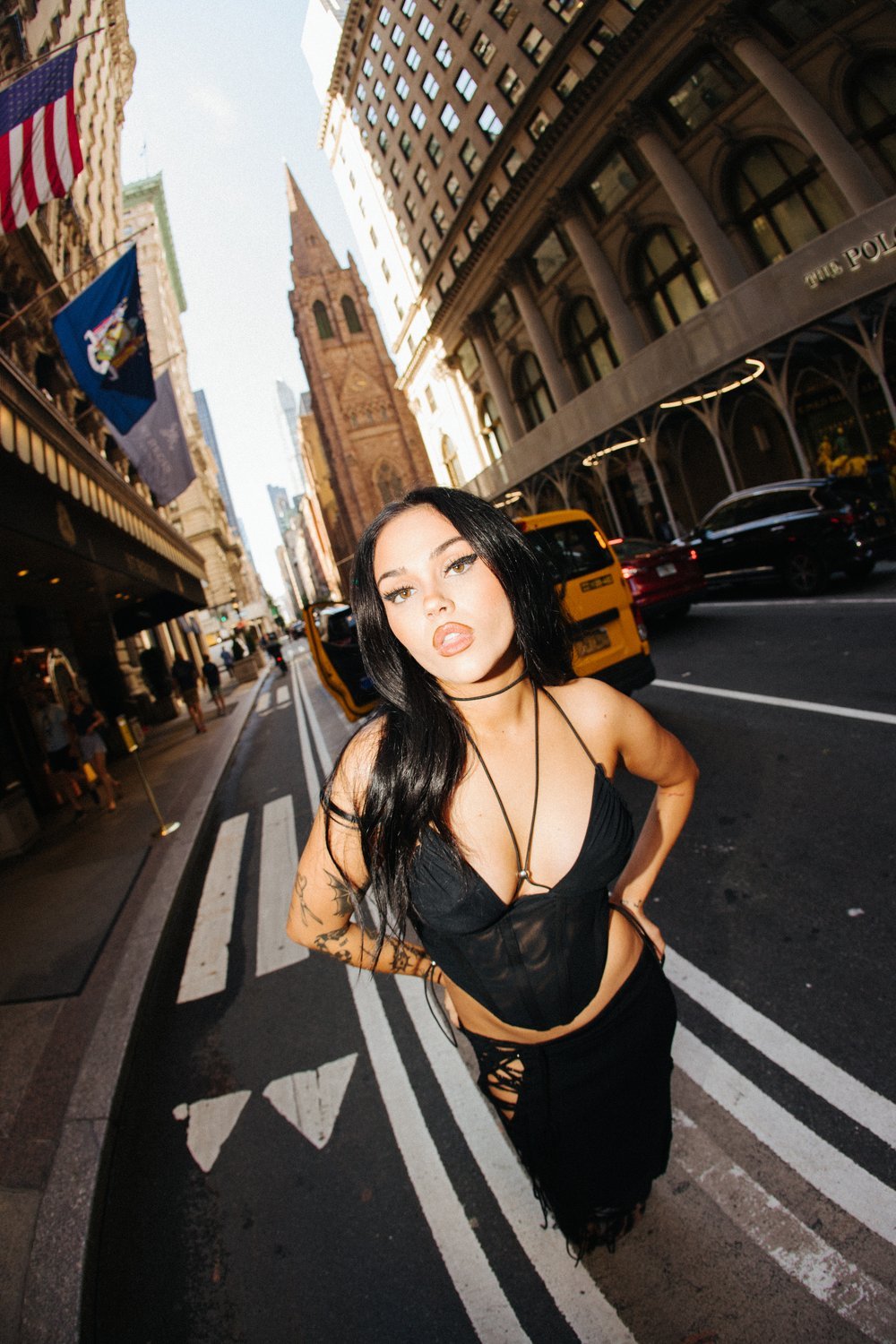Low-angle view of a young woman posing on a city street wearing a black top and skirt, with buildings and a yellow taxi in the background.