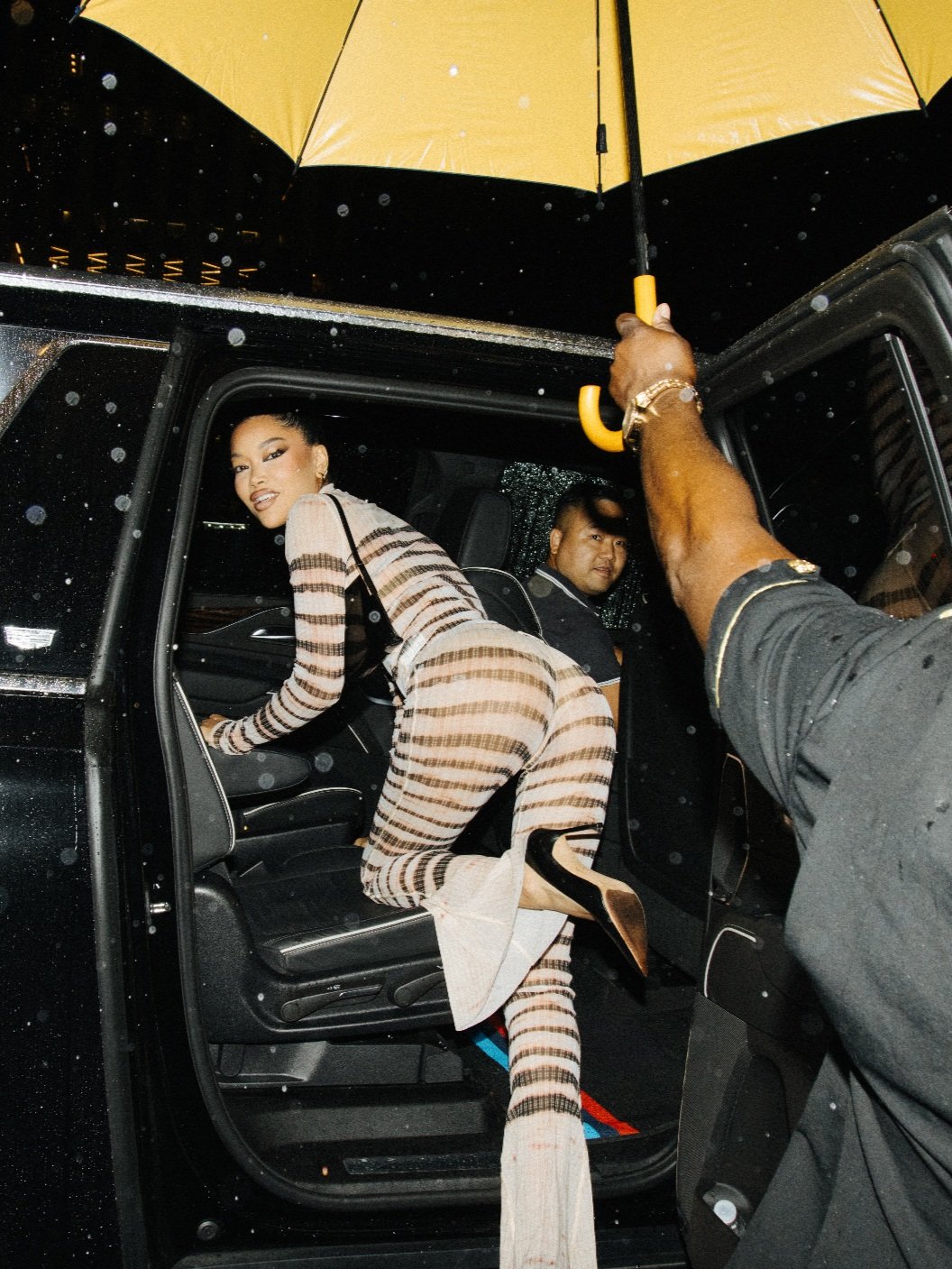 Adult female getting out of a black car, wearing a striped, long-sleeved jumpsuit and high heels, on a wet city street at night.