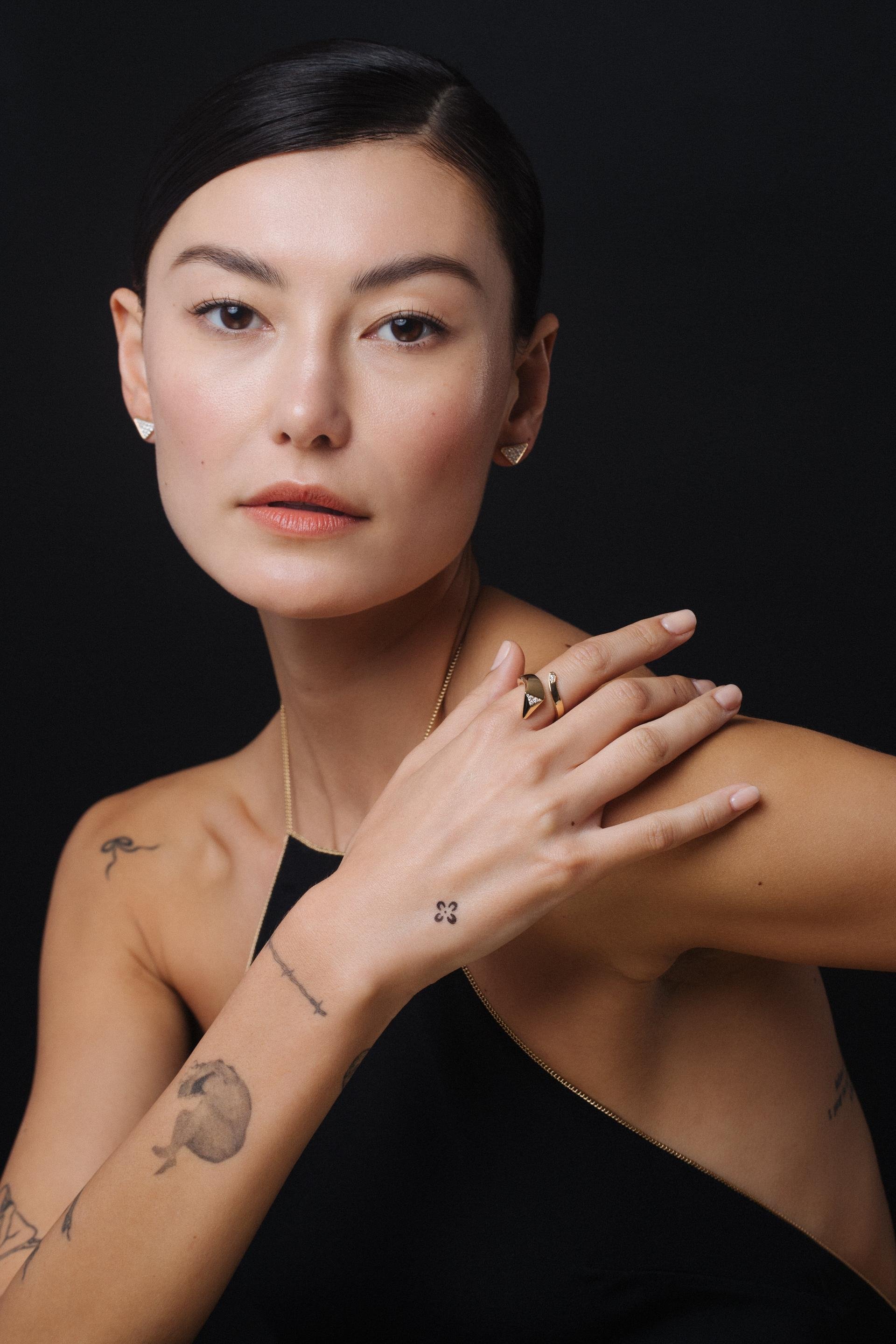 Close-up studio shot of a woman with a ring, earrings, and tattoos, against a dark background, emphasizing jewelry and personal style.