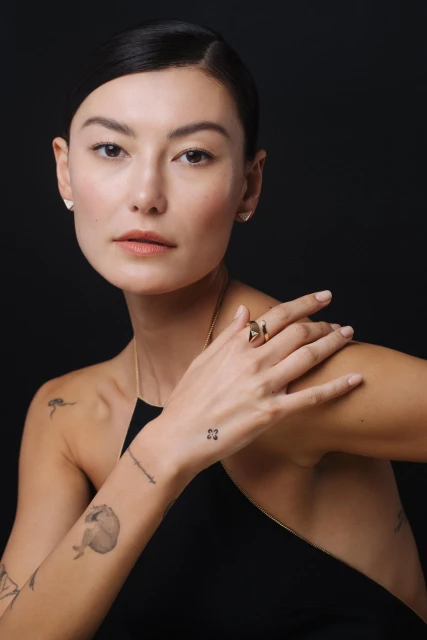 Close-up studio shot of a woman with a ring, earrings, and tattoos, against a dark background, emphasizing jewelry and personal style.