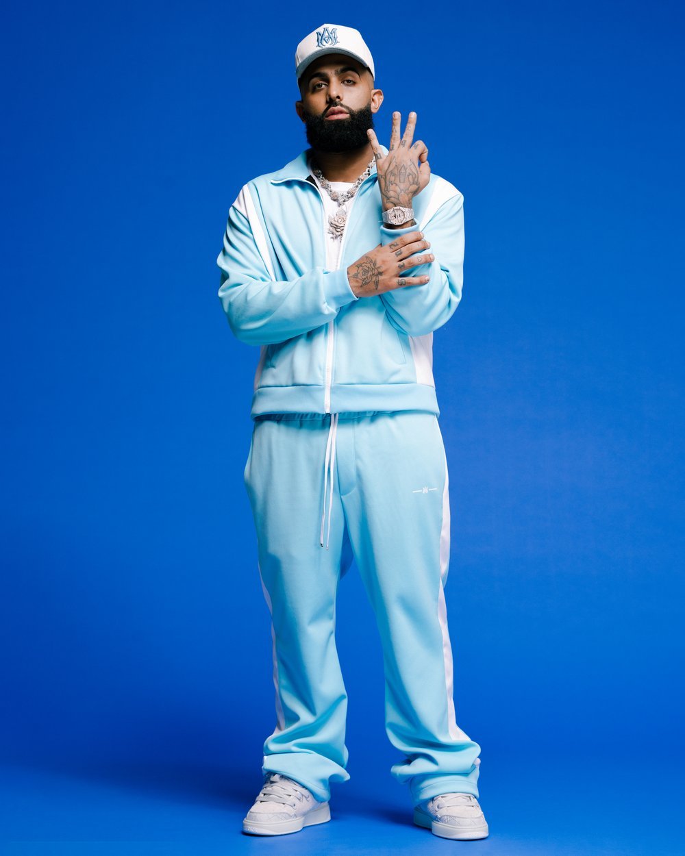 Full body shot of a man in a blue tracksuit, white sneakers, and baseball cap against a blue background, gesturing with his fingers.