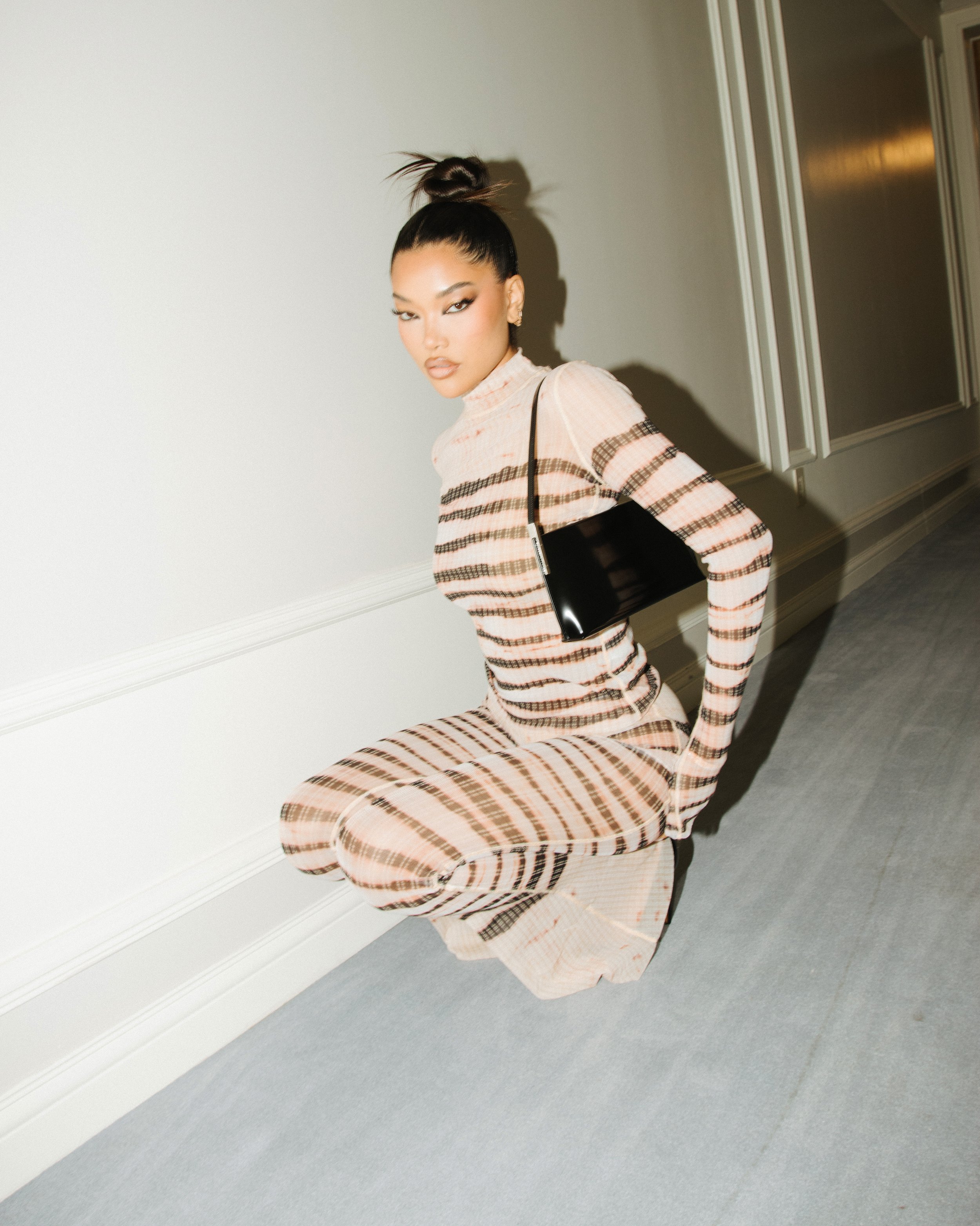 Elegant woman in striped dress poses crouching indoors, carrying a black purse. Fashion editorial portrait with neutral colors and mood.