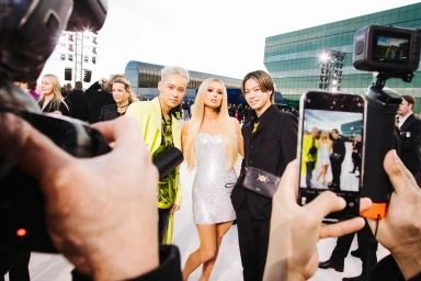 Paris Hilton posing with two young men surrounded by cameras and phones being held up in front of a modern building exterior.