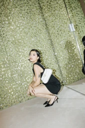 Woman in a black dress squatting against a textured green wall, holding a white shoulder bag.