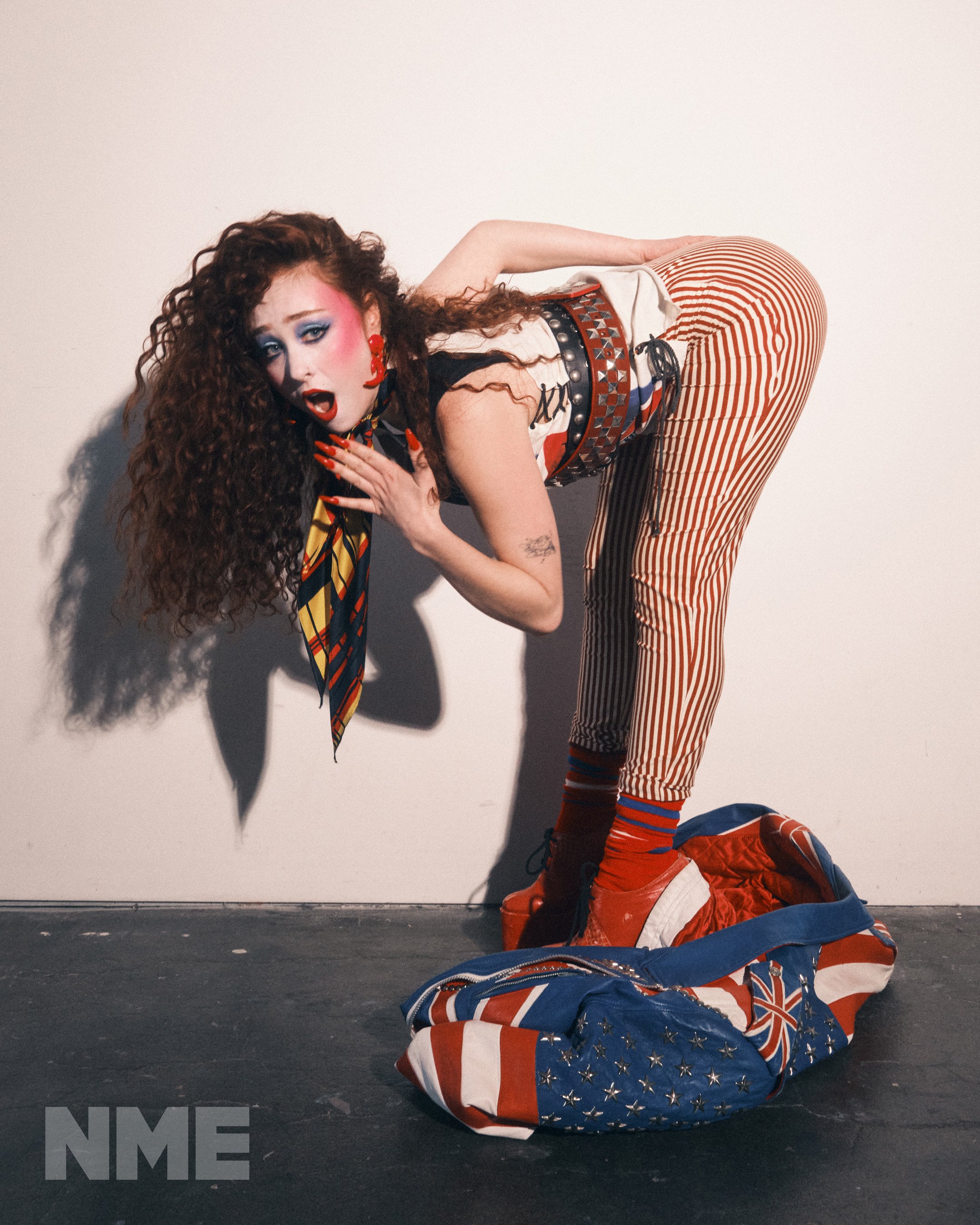 Young woman in a retro fashion pose, vibrant makeup, bold patterns, and red platform boots in a studio setting with a travel bag.
