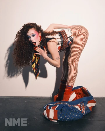 Young woman in a retro fashion pose, vibrant makeup, bold patterns, and red platform boots in a studio setting with a travel bag.