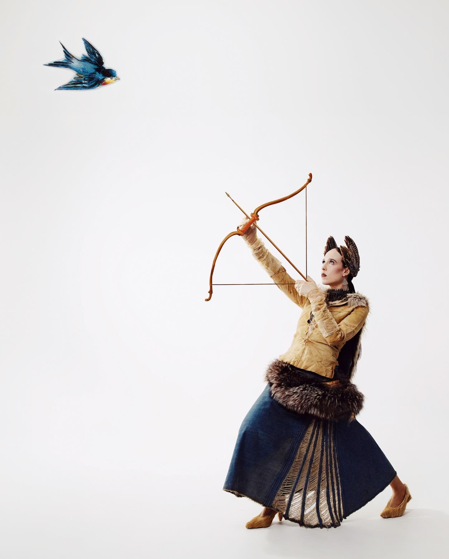 Woman archer aiming bow and arrow at blue bird in a studio shot against white backdrop. She's wearing fur-trimmed fashion and gold heels.