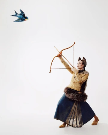 Woman archer aiming bow and arrow at blue bird in a studio shot against white backdrop. She's wearing fur-trimmed fashion and gold heels.