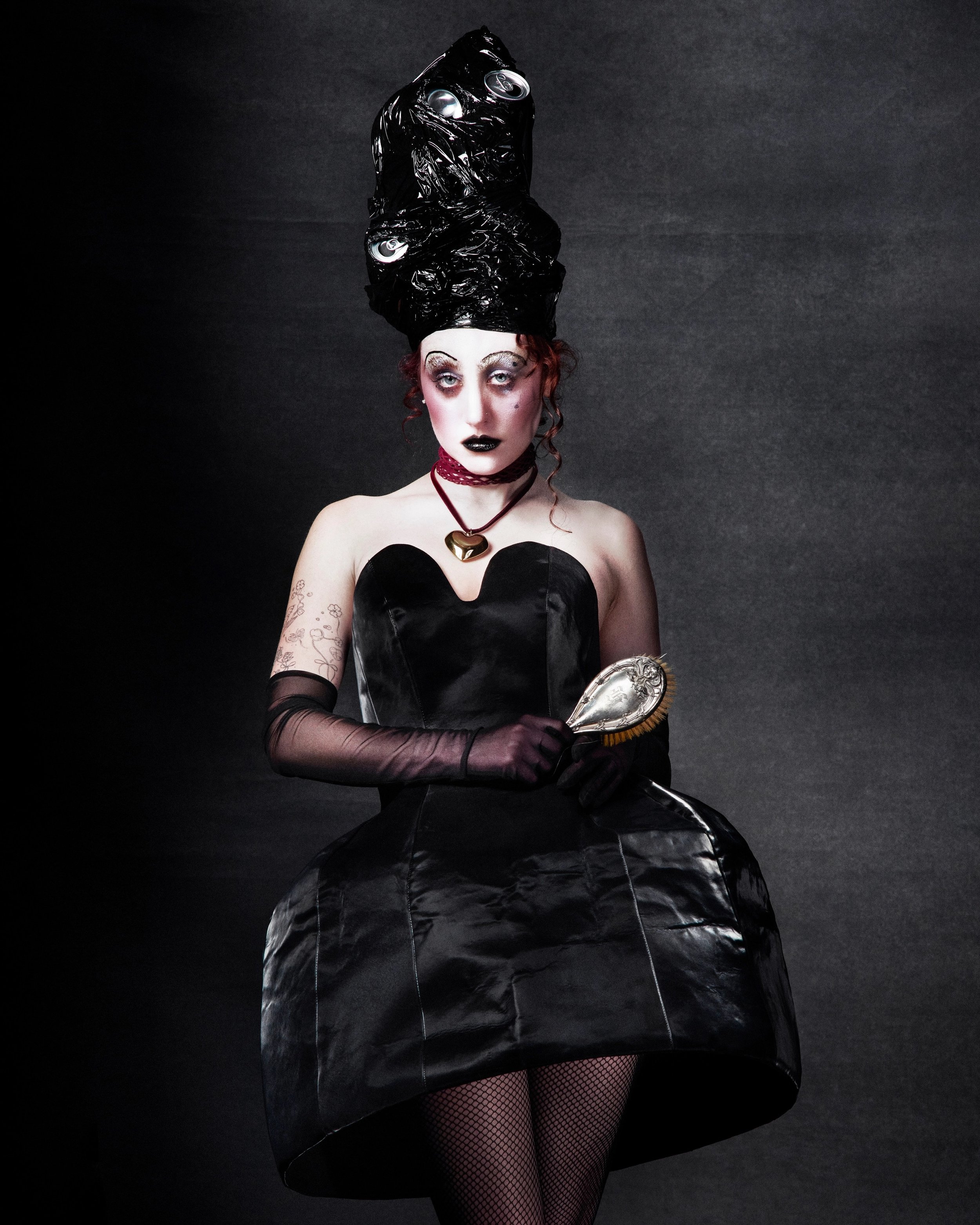 Portrait of a person with exaggerated makeup, wearing a black dress and elaborate black headpiece with reflective accents, holding a hairbrush.